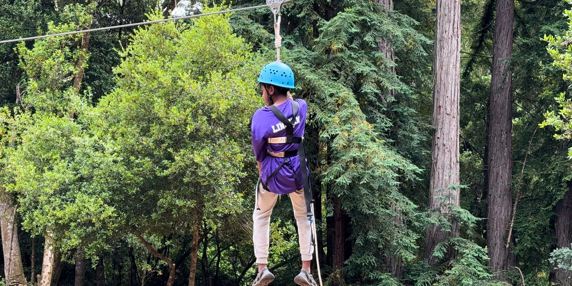 A Camp Loma Mar camper rides the zip line with their back to the camera in a blue hoodie.