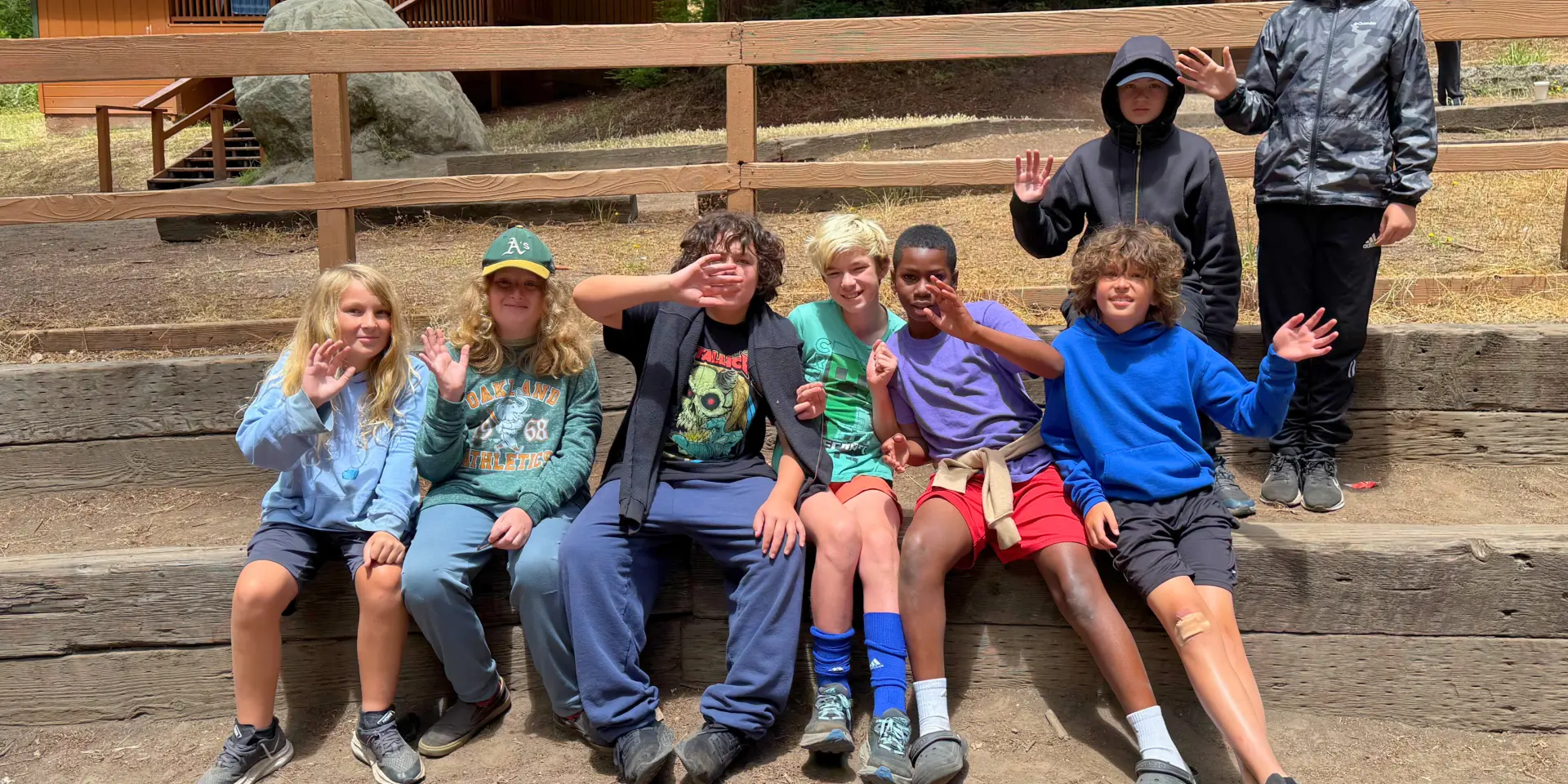 8 campers smile and wave at the camera while sitting on stepped benches in a recreation area in the woods at Camp Loma Mar.