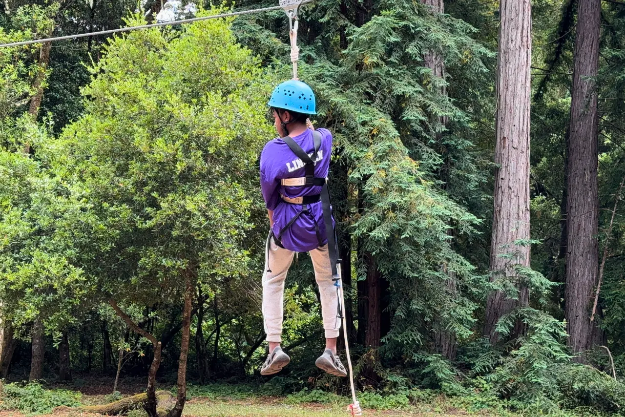 A Camp Loma Mar camper rides the zip line with their back to the camera in a blue hoodie.