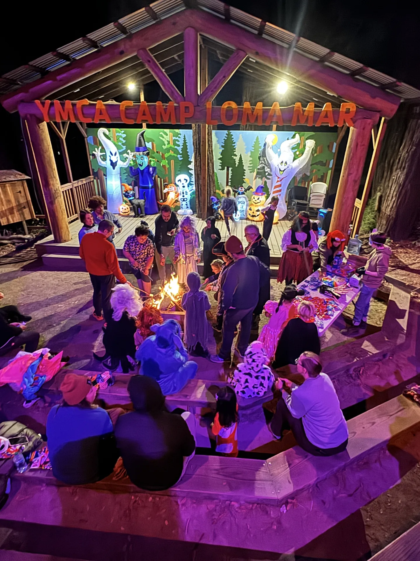 Adults and children sit and stand around a stage at Camp Loma Mar decorated for Halloween with inflatable characters and purple and orange lights.