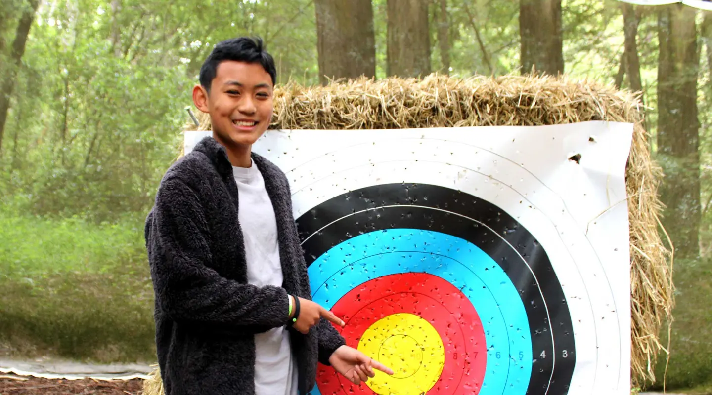 Camp Loma Mar male camper smiling and pointing at the archery target with bullseye