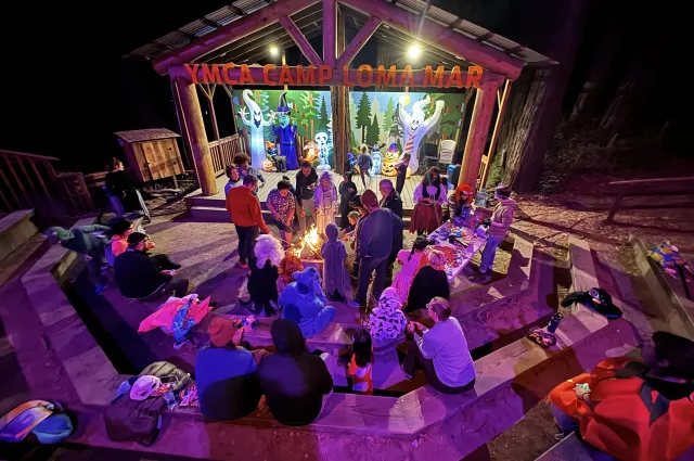Adults and children sit and stand around a stage at Camp Loma Mar decorated for Halloween with inflatable characters and purple and orange lights.