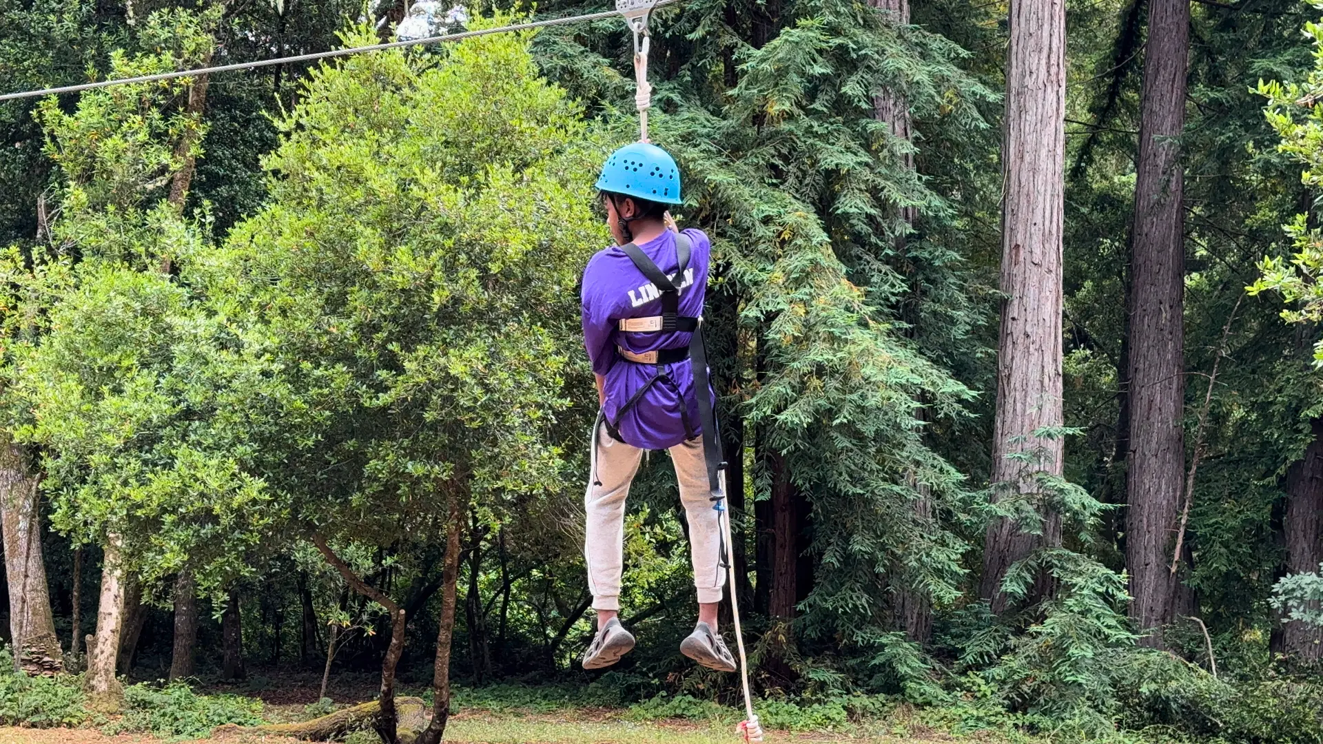 A Camp Loma Mar camper rides the zip line with their back to the camera in a blue hoodie.