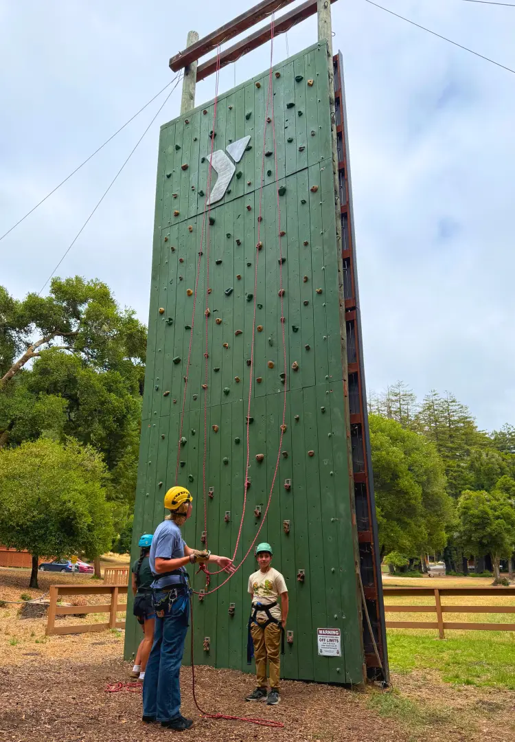A Camp Loma Mar counselor helps a camper put their gear on to climb the Camp Loma Mar climbing wall in a field of green grass.