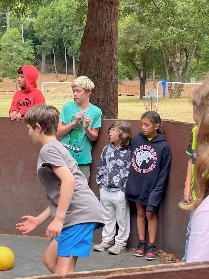 One child bats away a Gaga Ball in the Gaga pit while three other children watch from the sideline.