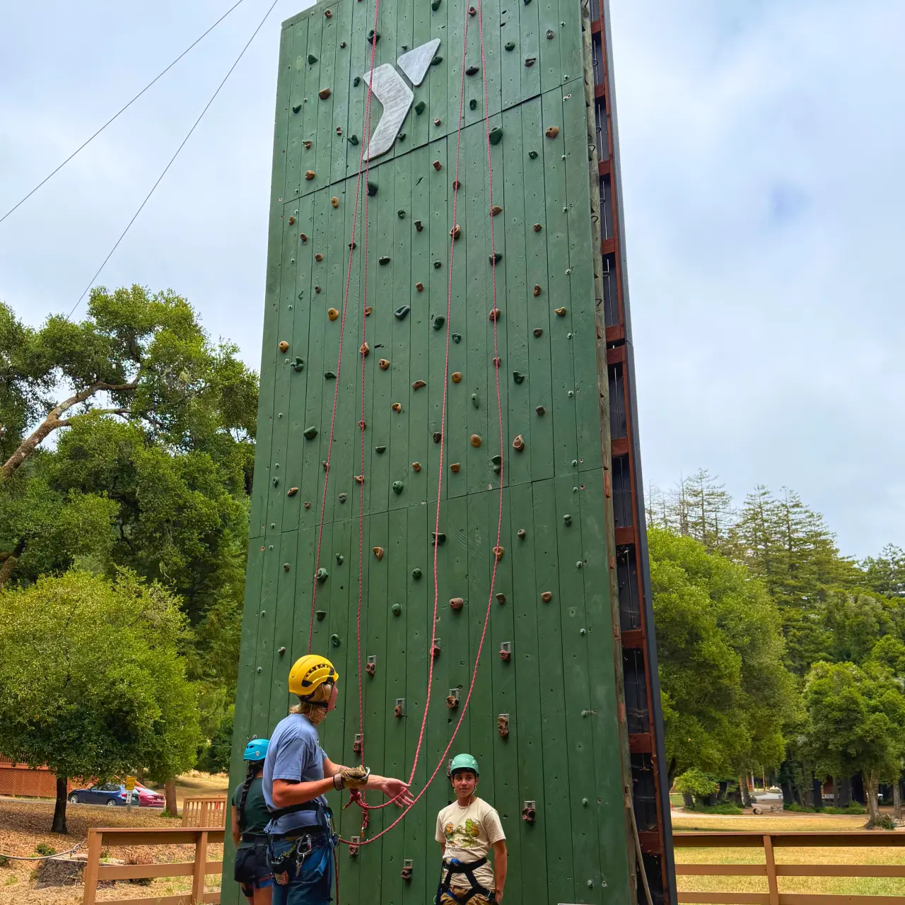 A Camp Loma Mar counselor helps a camper put their gear on to climb the Camp Loma Mar climbing wall in a field of green grass.
