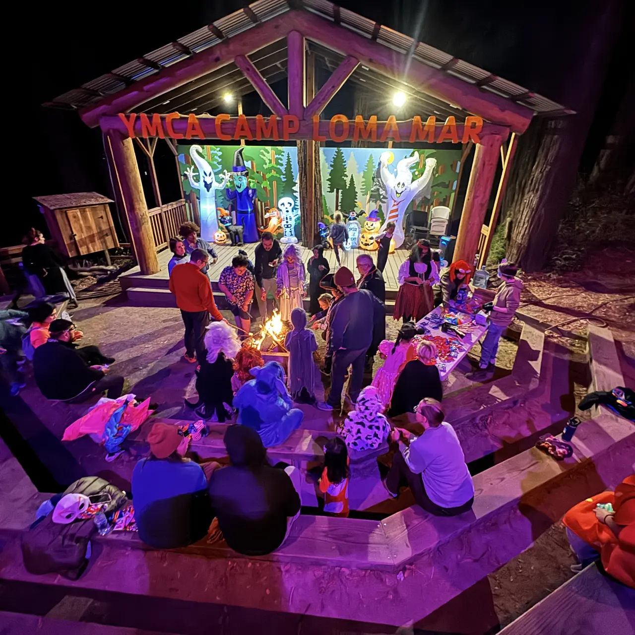 Adults and children sit and stand around a stage at Camp Loma Mar decorated for Halloween with inflatable characters and purple and orange lights.