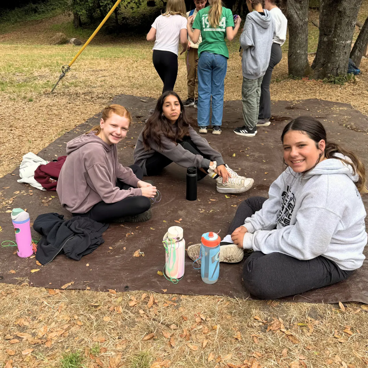 Three girls smile at the camera while sitting on a tarp in a field.