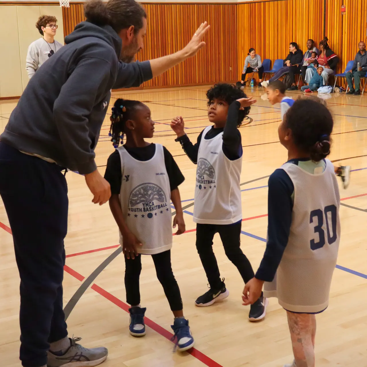 Coach holds up a high five to a basketball player in a group of 3 kids wearing white jerseys on a basketball court.
