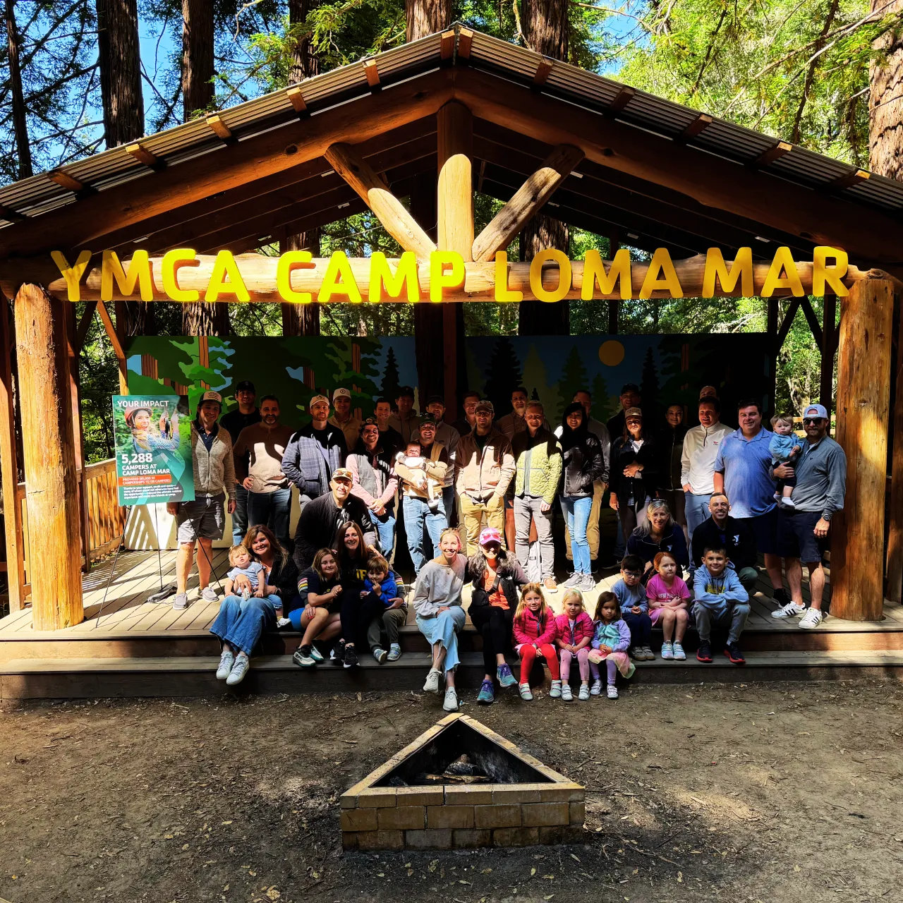 A group of adults and children standing on the Camp Loma Mar Campfire Bowl stage smiling and enjoying the redwood forest.