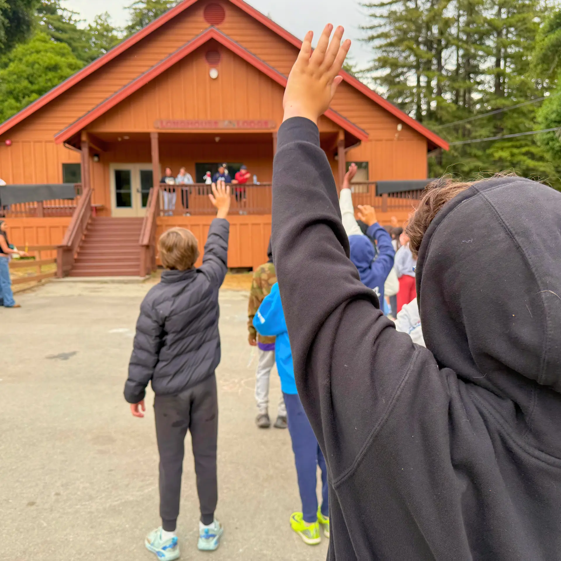 Two boys in black jackets raise their hands in the lunch line in front of the Camp Loma Mar lodge.