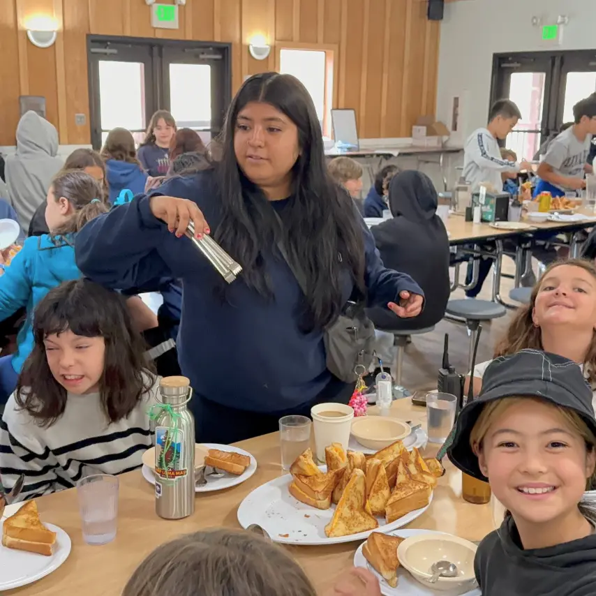 A counselor hands out food to campers in the Camp Loma Mar dining hall, a few of the children smile at the camera.